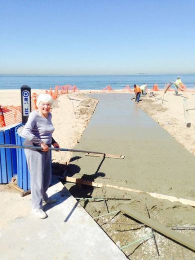 這張拍攝於海灘的照片中，一位白人老太太笑咪咪地拿著金屬管站在正在建造的無障礙步道旁，可見遠方正在施工的工作人員，以及背景一望無際的海與天。照片來源 Manhattan Beach Timeline 臉書粉絲頁。