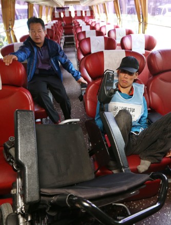 一名身障者坐在巴士內的椅子上，他的前方走道擺放著是傾斜的輪椅，可見走道與座位的狹小，無法讓他順利使用輪椅上車。斜後方做了另一位男士。身障者要上公車，需要司機協助。攝影 Yonhap