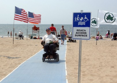 這張拍攝於沙灘上的照片，有一位電動輪椅使用者正在淡藍色的沙灘墊上步行，一旁可見美國國旗，令一旁是紐約市公園管理處的楓葉旗幟的標誌，背景可見海灘人潮與海。照片前景有一個樹立的標示，圖繪有海灘墊、輪椅使用者、家長與手推的娃娃車與小孩，標示文字是Beach Access Surface 可及海灘的鋪面步道。